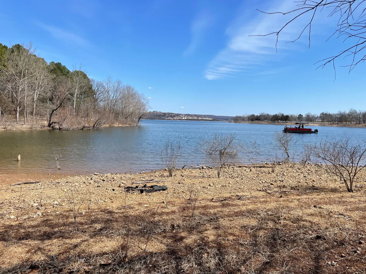 A wide view of the calm surface of Table Rock Lake, framed by bare trees and distant hills, with a small boat on the water. The image captures the tranquil atmosphere and outdoor activities like fishing, boating, and wildlife observation.