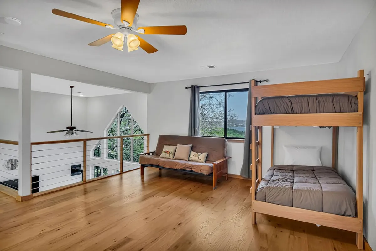 A spacious loft area with a twin bunk bed, a futon couch, large window, and wooden railing. The space is set up for sleepovers, reading, and relaxing, with plenty of natural light and a playful yet restful ambiance.