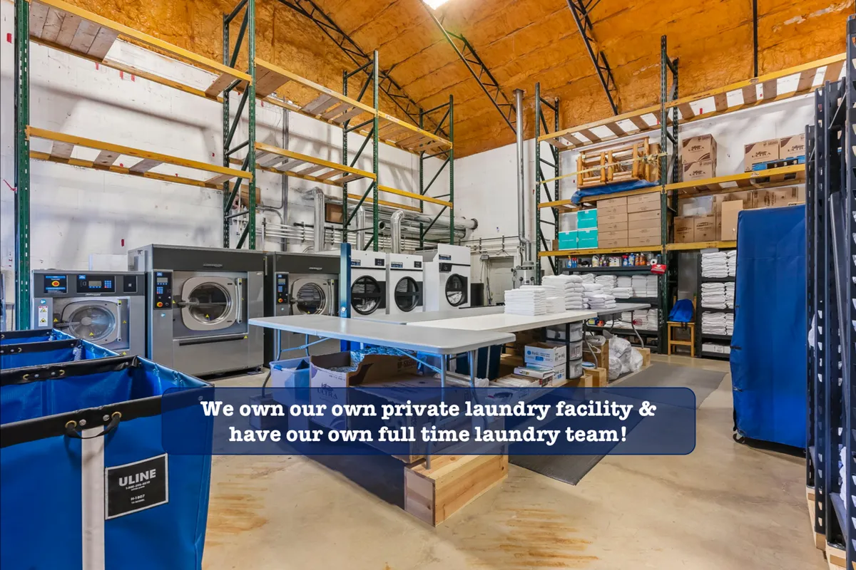 An organized commercial laundry facility with industrial washing machines, a large folding table, and laundry carts. The area is busy yet immaculate, reflecting professional standards for cleanliness and service.