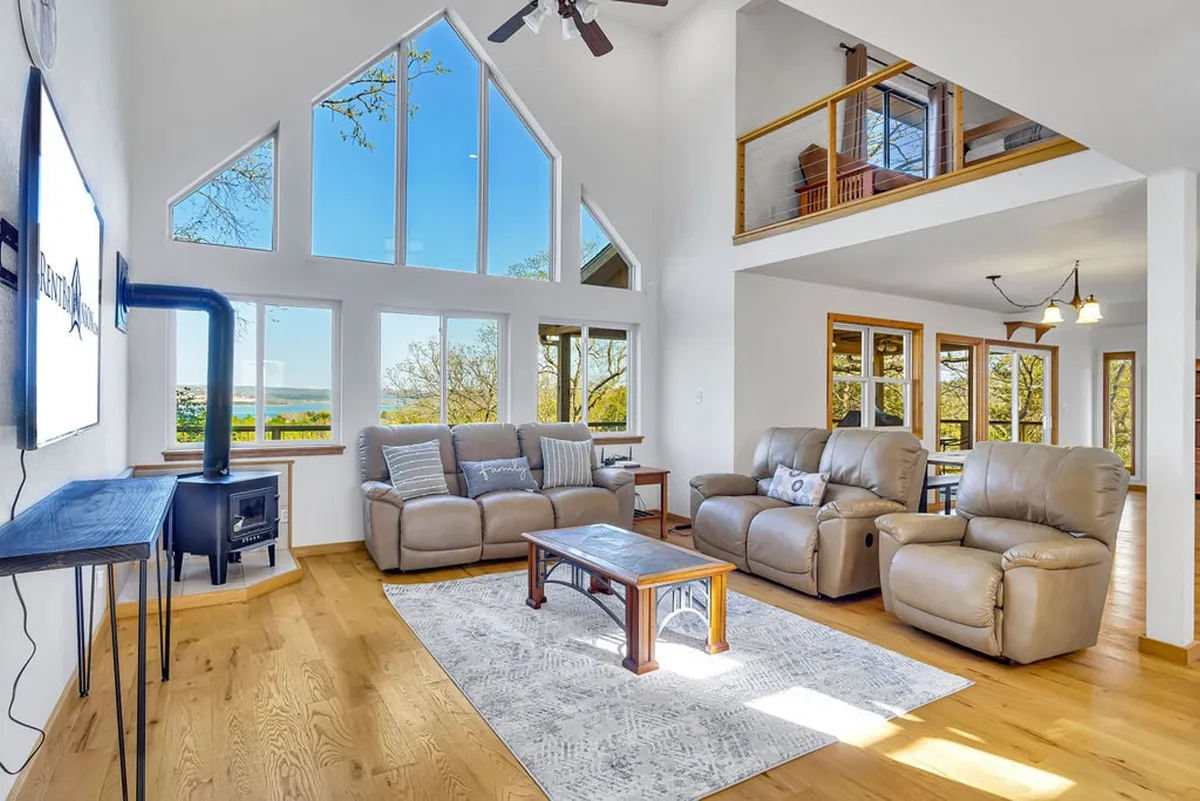 The image showcases a cozy living space with inviting, plush seating arranged around a warm fireplace. Expansive windows frame breathtaking views of Table Rock Lake, filling the room with natural light and offering a front-row seat to stunning sunsets. A wooden coffee table sits at the center, perfect for gathering or playing games, creating a welcoming setting for family and friends to relax and reconnect.